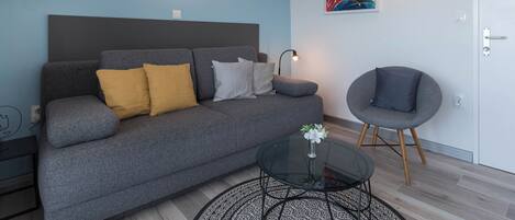 Uma sala de estar moderna com um sofá cinzento, uma mesa de centro redonda e uma poltrona cinzenta. Há um quadro na parede e um aparelho de ar condicionado fixado na parede.