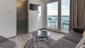 En moderne stue med et glasbord, en sofa, et fjernsyn ophængt på væggen og havudsigt gennem store vinduer.