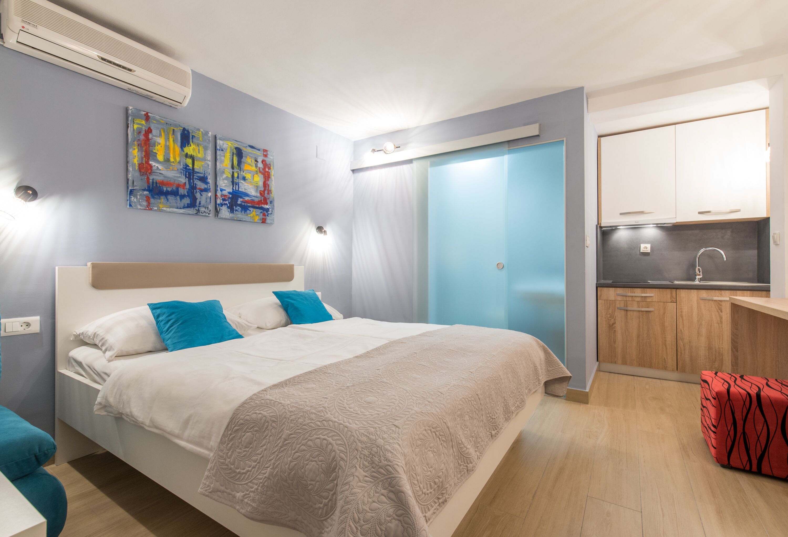 A modern bedroom with a bed, a small kitchenette, and a bathroom with a blue door.