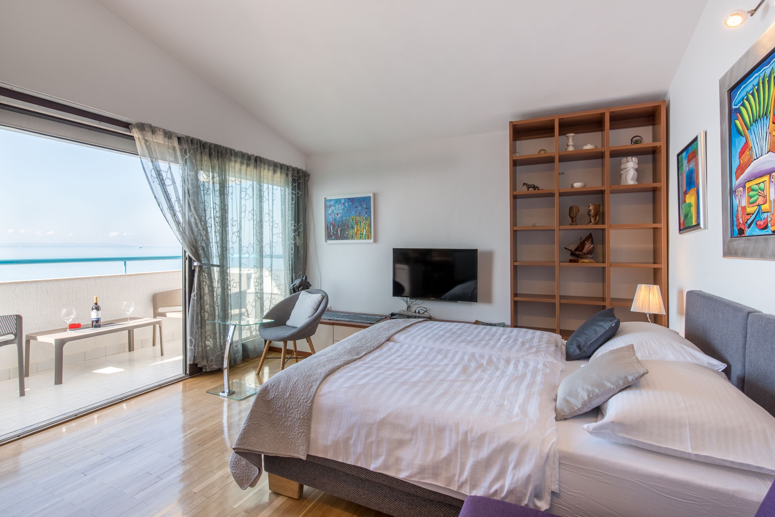 A modern bedroom with a large bed, a TV, a bookshelf, and a balcony with a view of the sea.