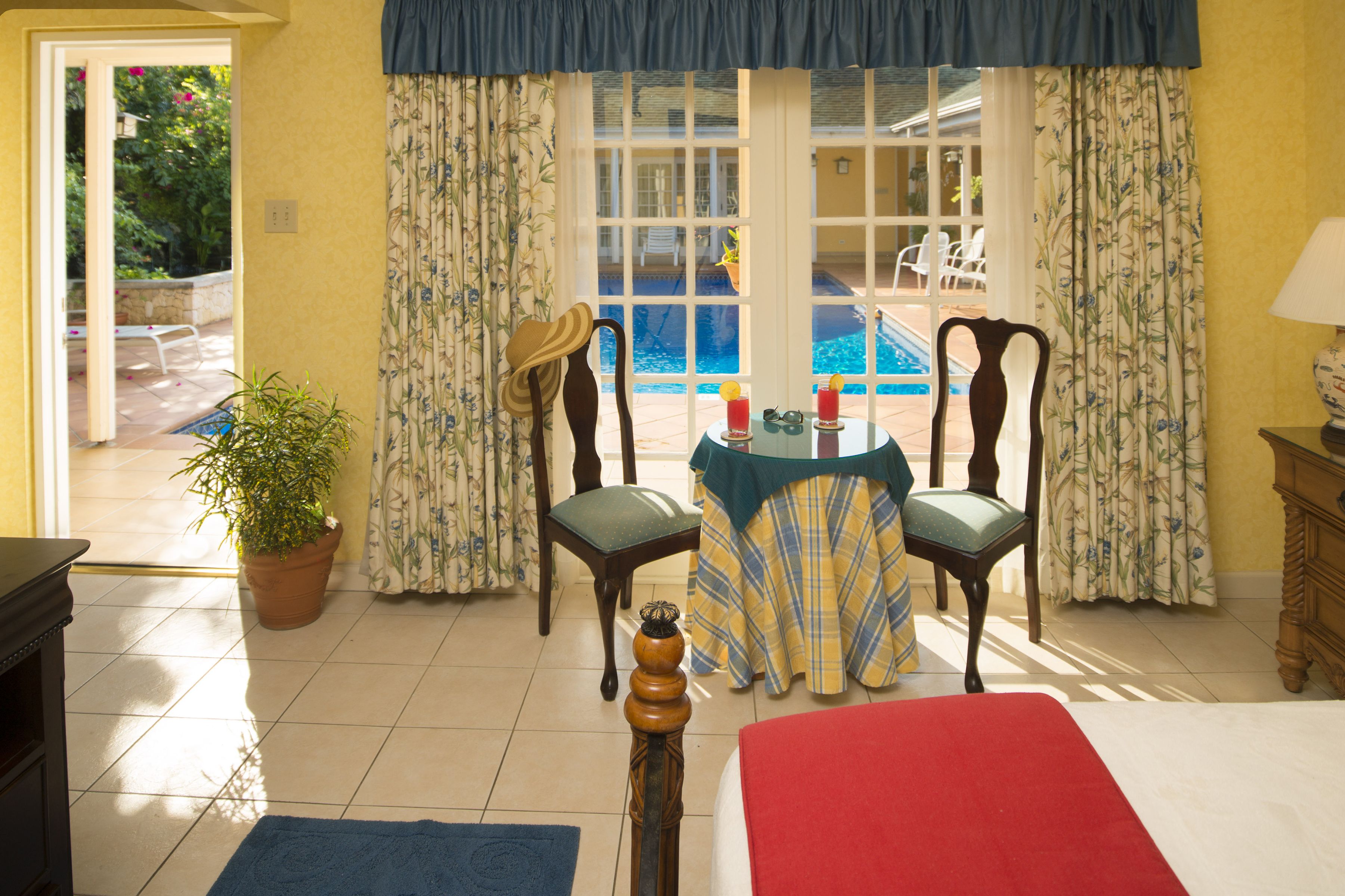 A room with a bed, a table with two chairs, a potted plant, and a view of a swimming pool through large windows.