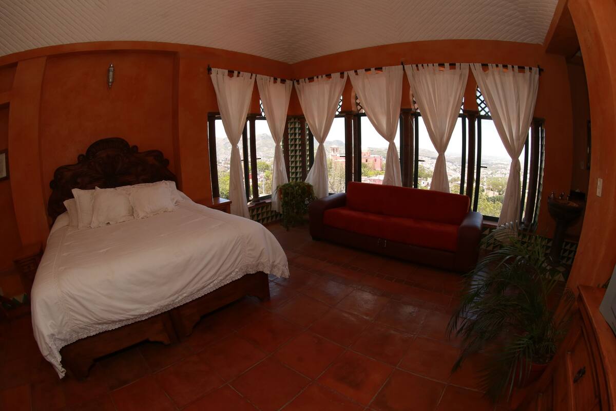 A bedroom with a bed, a red sofa, and large windows with curtains.