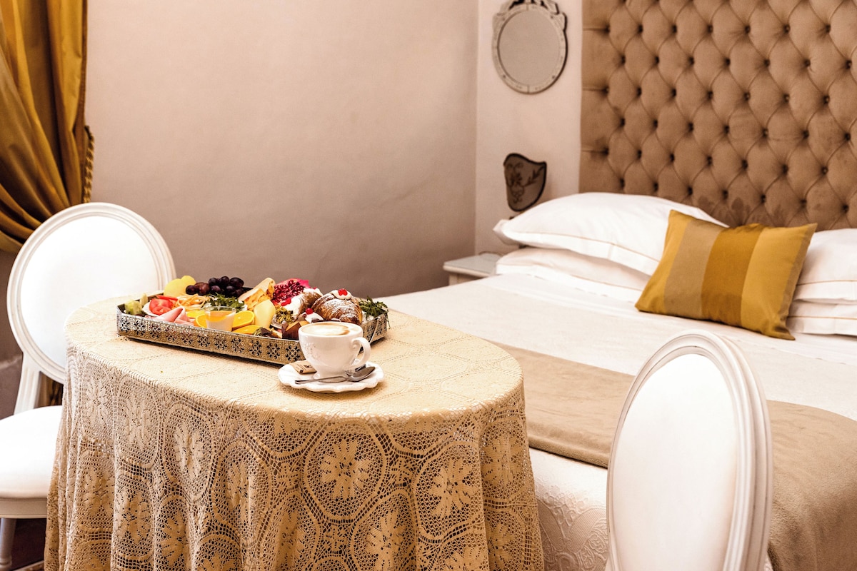 Una mesa de desayuno bien dispuesta con una bandeja de frutas variadas, una taza de café y un plato decorativo. Se ve una cama con cabecera capitonada y una almohada amarilla en el fondo.
