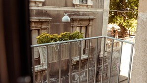Un balcón con cestas de mimbre que contienen flores amarillas, una barandilla de metal y un edificio con ventanas ornamentadas al fondo.