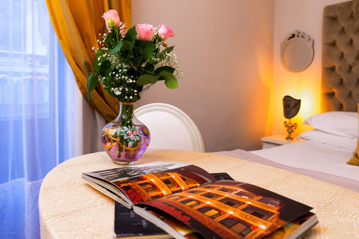 A hotel room with a bed, a nightstand with a lamp, a vase with flowers, and a magazine on the table.