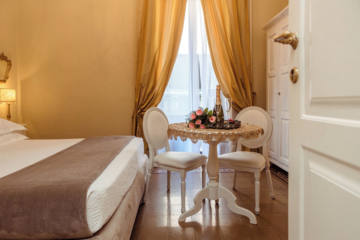 A hotel room with a bed, a round table with a floral centerpiece, two chairs, and a door with a gold handle.