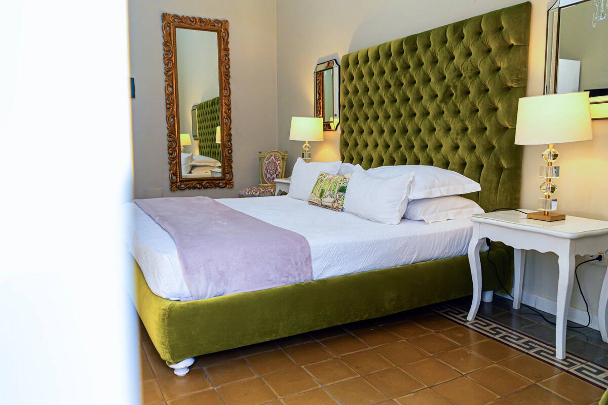 A hotel room with a green upholstered bed, white linens, a large mirror, two bedside lamps, and a small white nightstand.