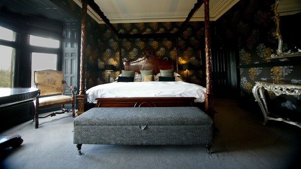 A four-poster canopy bed with a footboard, a bench, and a chair in a room with floral wallpaper and a large window. - Mitton Hall Country House Hotel (Clitheroe)