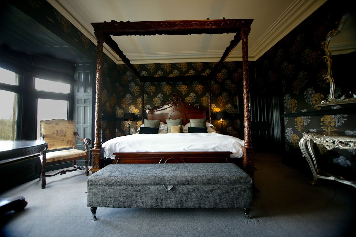 A four-poster canopy bed with a footboard, a bench, and a chair in a room with floral wallpaper and a large window.