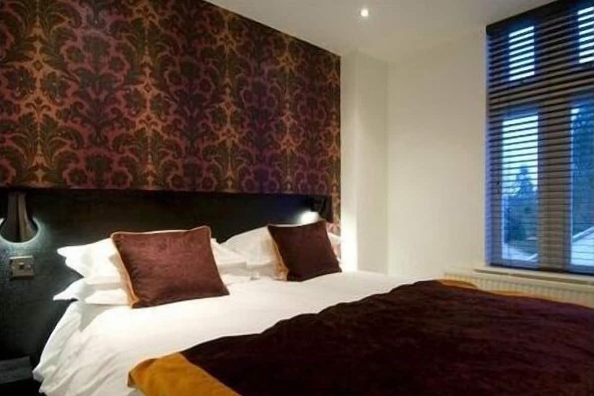 A hotel room with a patterned wallpaper, a bed with white linens and two brown pillows, and a window with blinds.
