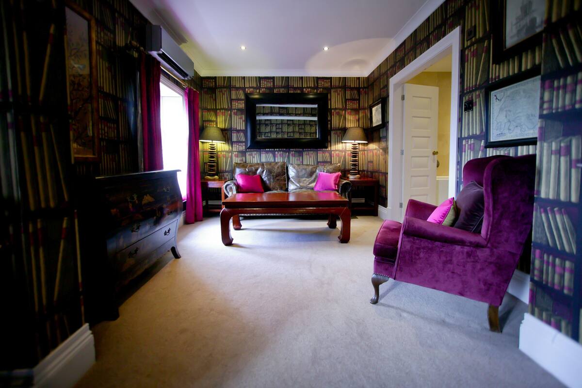 A room with a large wooden coffee table, a purple armchair, and a bookshelf filled with books.