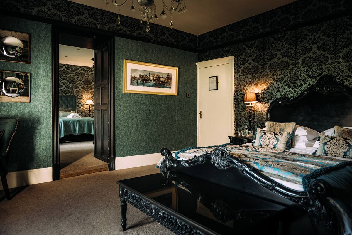 A bedroom with a large bed, a desk, and a chair. There is a door leading to another room.