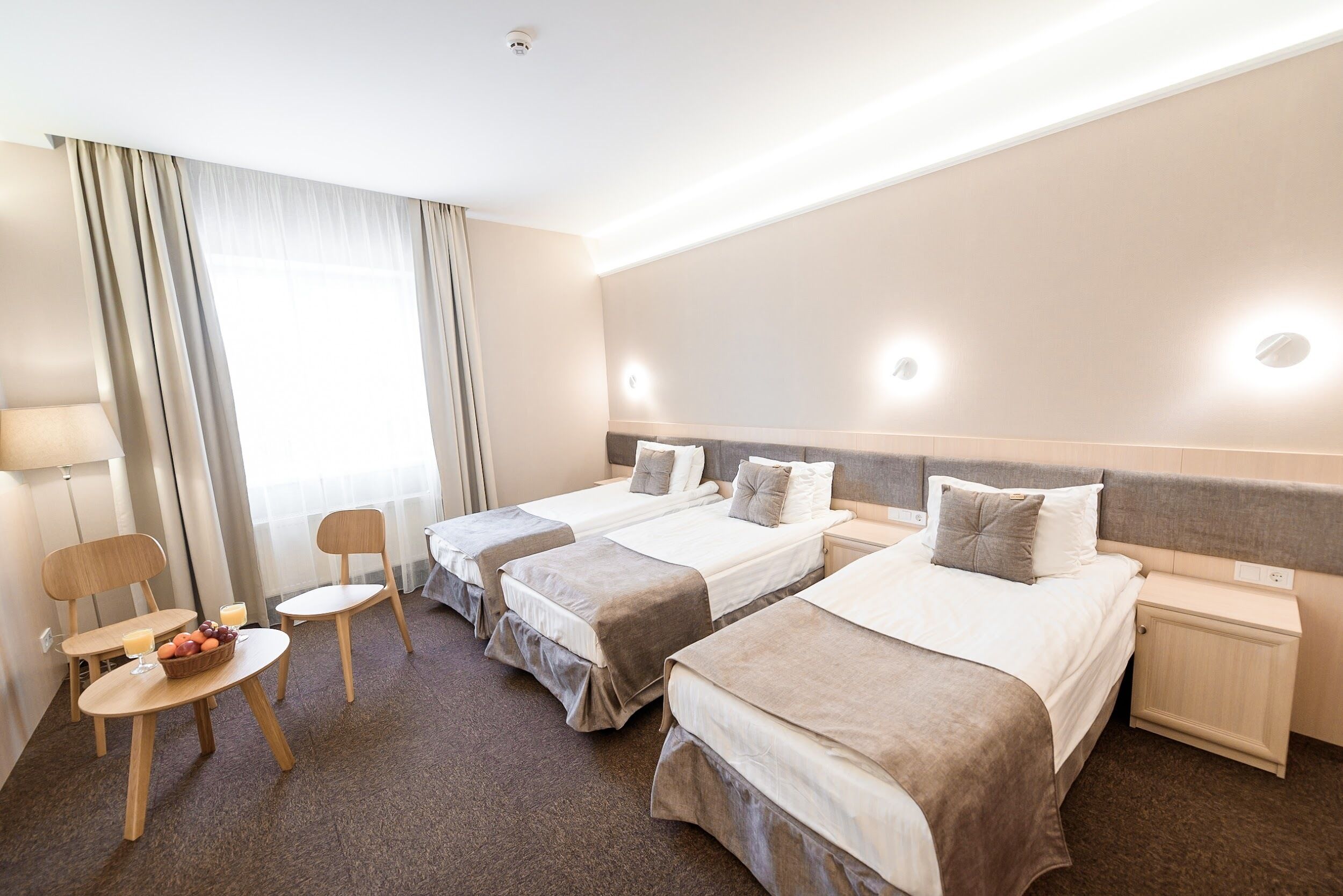 Une chambre d’hôtel avec trois lits simples, une petite table sur laquelle se trouve un panier de fruits, une chaise et une table de chevet avec une lampe.