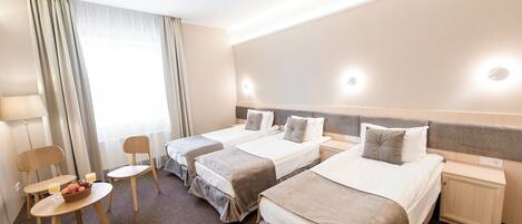 A hotel room with three single beds, a small table with a fruit bowl, a chair, and a bedside table with a lamp.