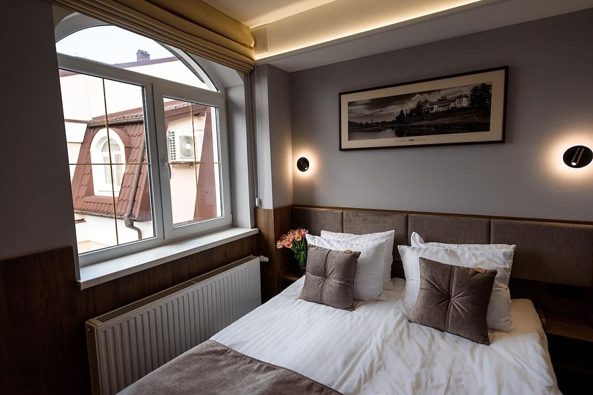 Ein Hotelzimmer mit großem Fenster, einem gerahmten Bild an der Wand und einem Bett mit vielen Kissen.