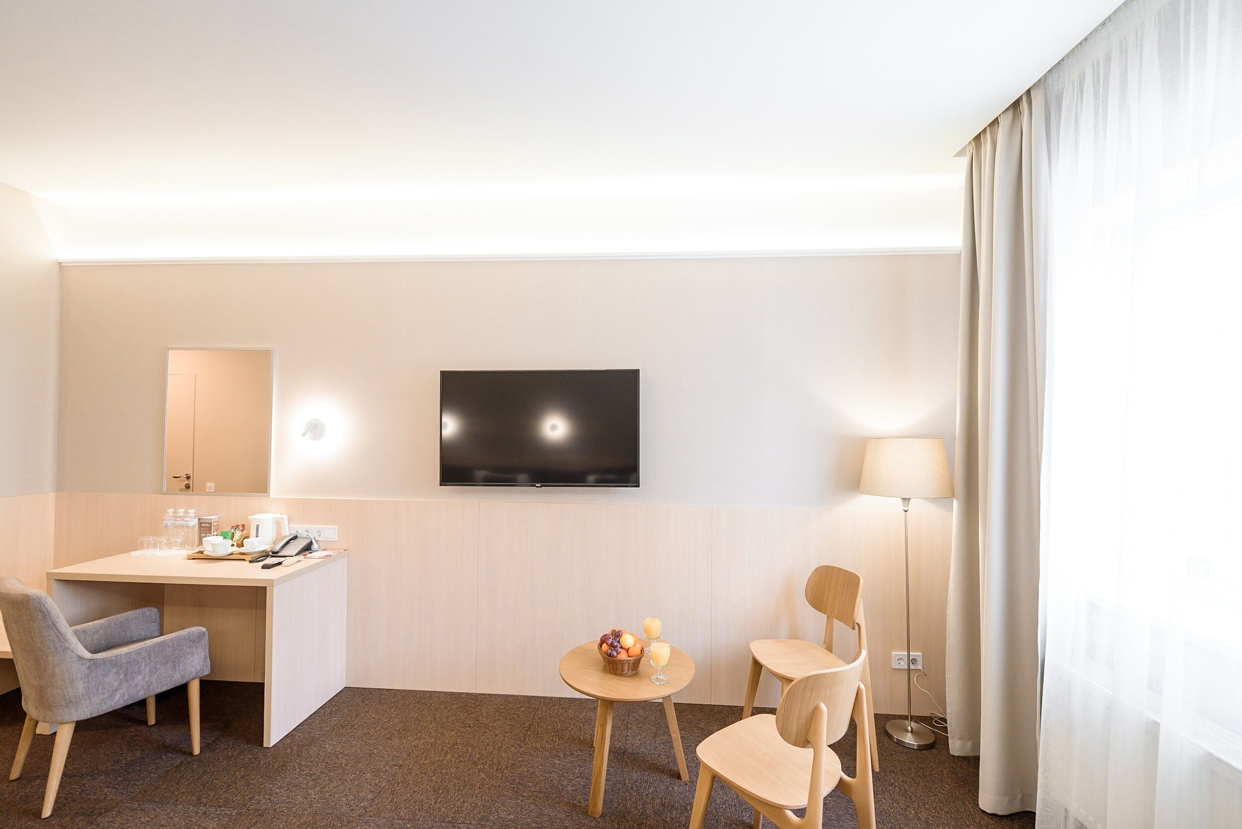 A hotel room with a desk, two chairs, a TV, a lamp, and a small table with fruit.