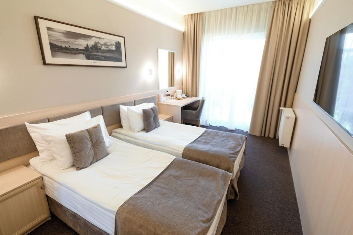Ein Hotelzimmer mit zwei Betten, einem Schreibtisch, einem Stuhl, einem großen Fenster mit Vorhängen, einem gerahmten Bild an der Wand und einem Fernseher.