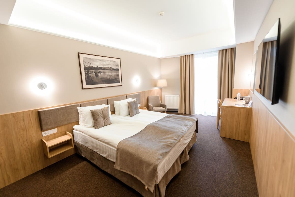 Ein Hotelzimmer mit einem großen Bett, Nachttischen, einem Schreibtisch, einem Sessel, einer Lampe, einem Fernseher und einem Fenster mit Vorhängen.