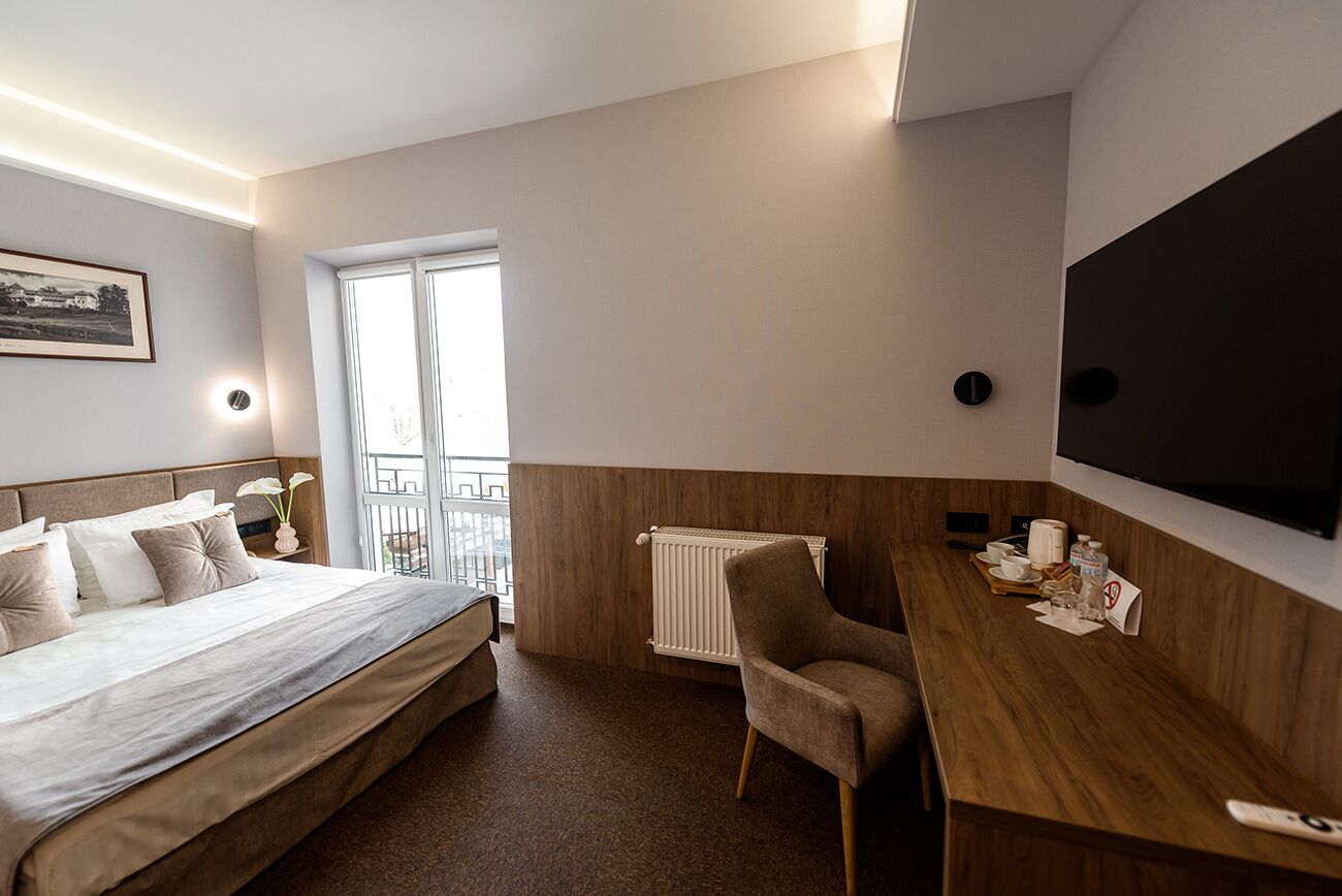 A hotel room with a bed, a desk, a chair, a radiator, a TV, and a window with a view.