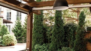 An outdoor patio with a glass-top table and wicker chairs, surrounded by greenery and a wooden pergola. - Nota Bene Hotel (Lviv)