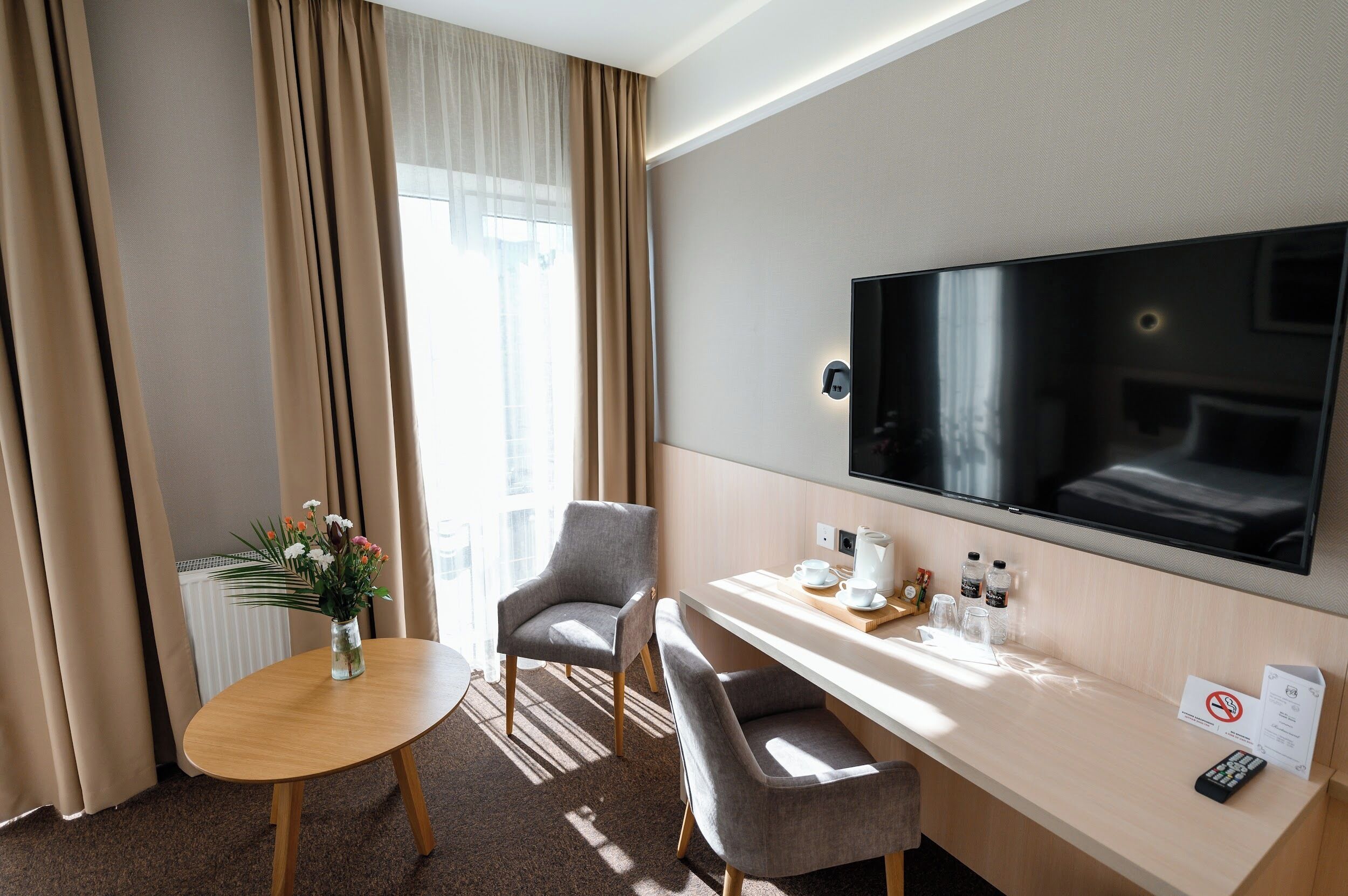 Habitación de hotel con cama, televisión, escritorio con silla, una mesita con un jarrón de flores y una ventana con cortinas.