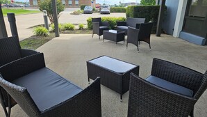 Un área de estar al aire libre con mobiliario de mimbre y una pequeña mesa, rodeada por setos y un edificio al fondo.