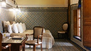 A room with a patterned tile floor, a bed with a striped bedspread, a wooden chair, a small table with a book, and a wall-mounted air conditioning unit. - Dar El Bali (Fes)