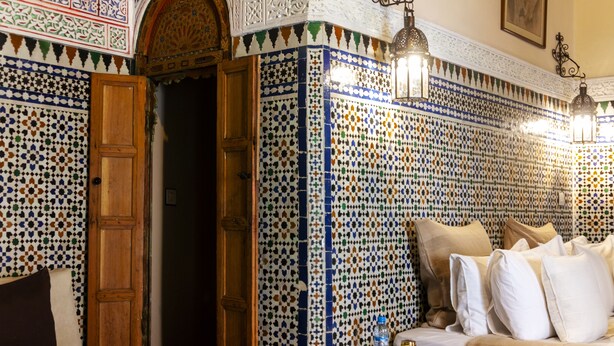 A room with a patterned tile wall, a wooden door, a sofa with pillows, and a small table with a bottle. - Dar El Bali (Fes)