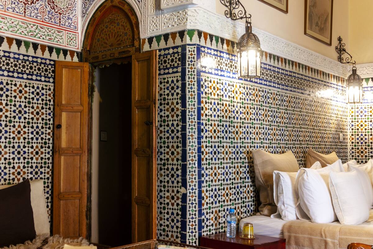A room with a patterned tile wall, a wooden door, a sofa with pillows, and a small table with a bottle.