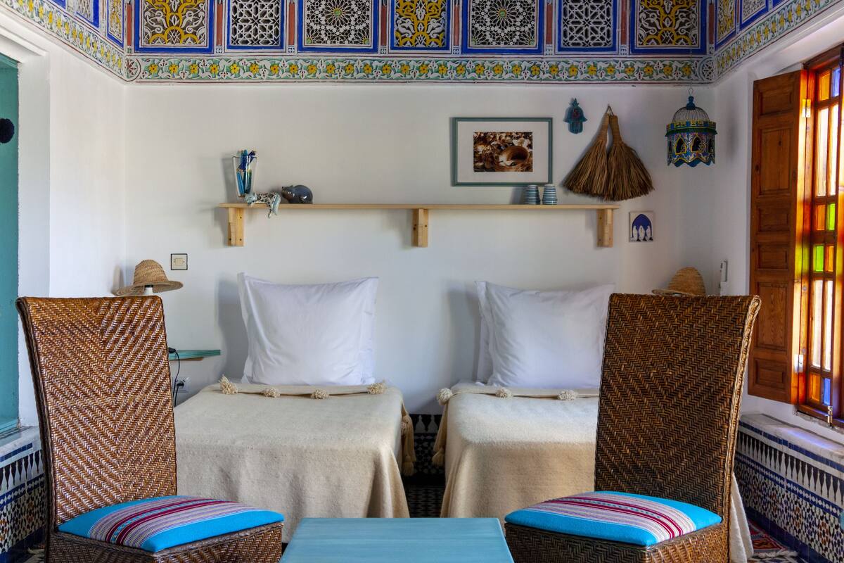 A room with two beds, wicker chairs, a small table, and a decorative ceiling.