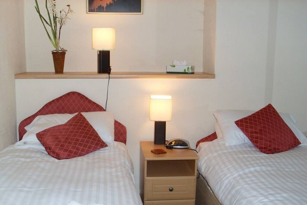 A hotel room with two beds, each with red pillows and white bedding. There are bedside tables with lamps and a small shelf above the beds.