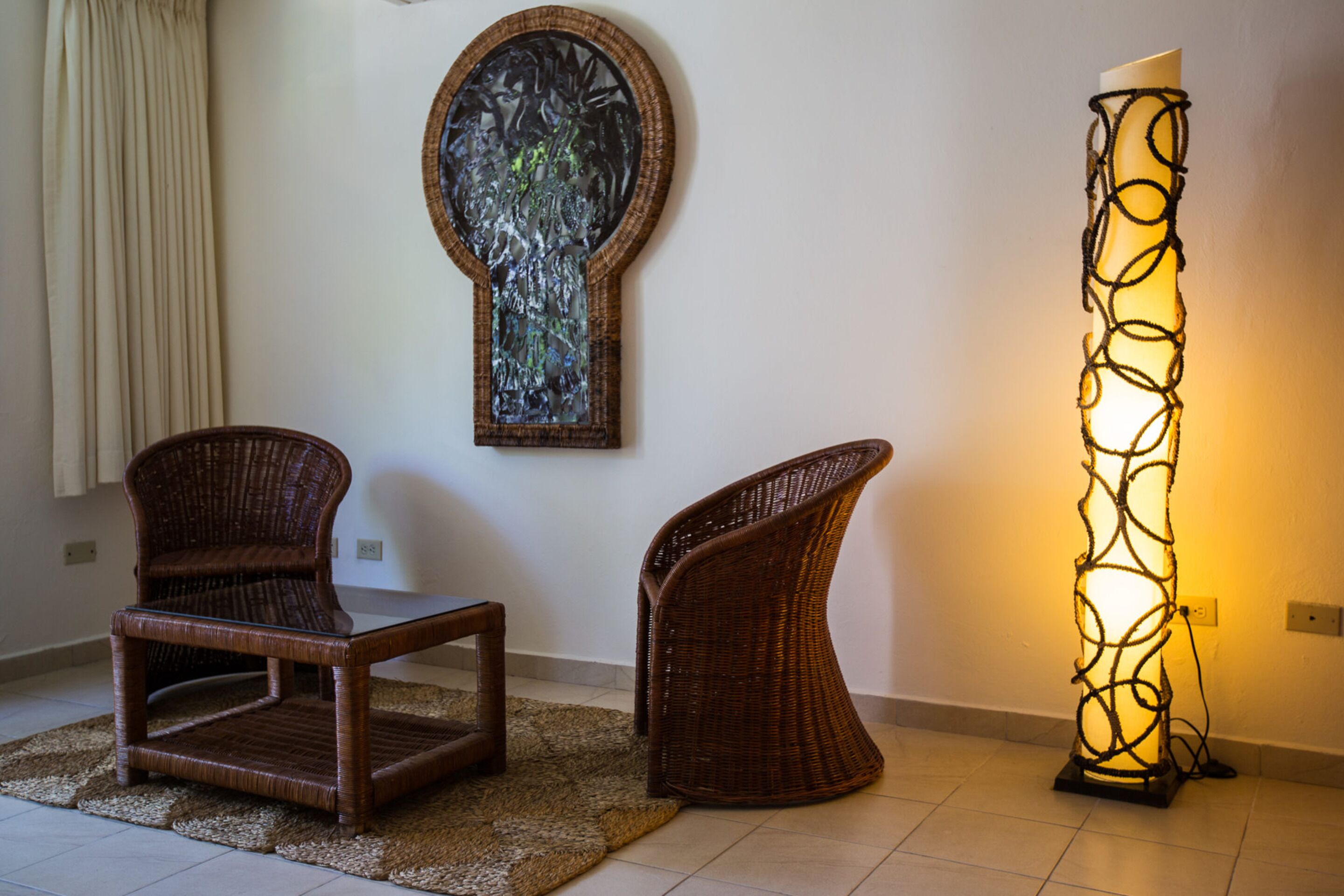 Una habitación con dos sillones de mimbre, una lámpara de pie tejida y una obra de arte abstracta colgada en la pared.