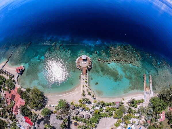 An aerial view of a resort with a central circular building surrounded by a beach and clear waters.