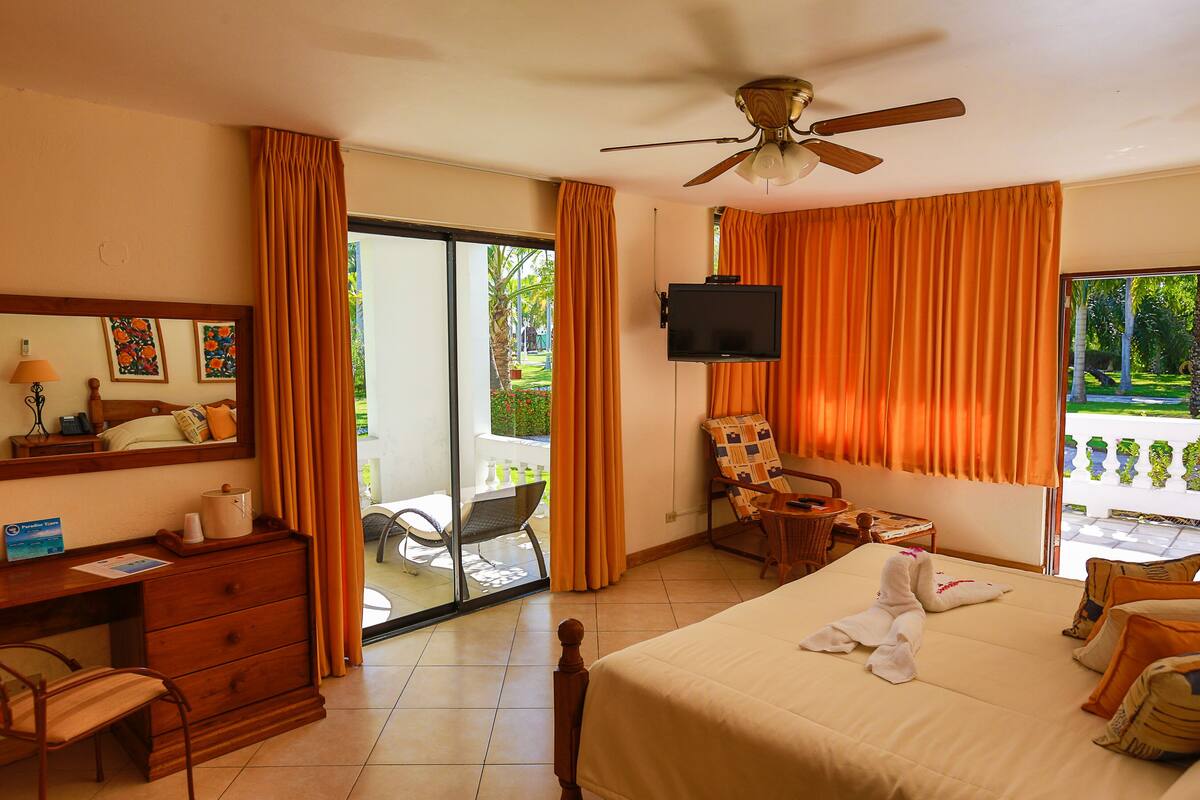 Une chambre d’hôtel avec un lit, un bureau, une chaise, une télévision et un balcon agrémenté de rideaux orange.
