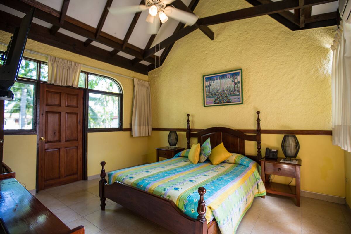 Une chambre avec un lit en bois, un ventilateur de plafond, une table de chevet, une fenêtre avec des rideaux et un tableau au mur.