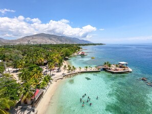 A tropical beach resort with clear turquoise waters, palm trees, and a mountainous backdrop.