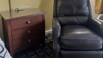 A hotel room with a bed, a brown nightstand with drawers, a black leather chair, and two floor lamps.