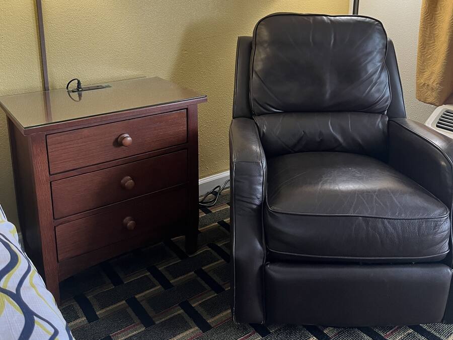 A hotel room with a bed, a brown nightstand with drawers, a black leather chair, and two floor lamps.