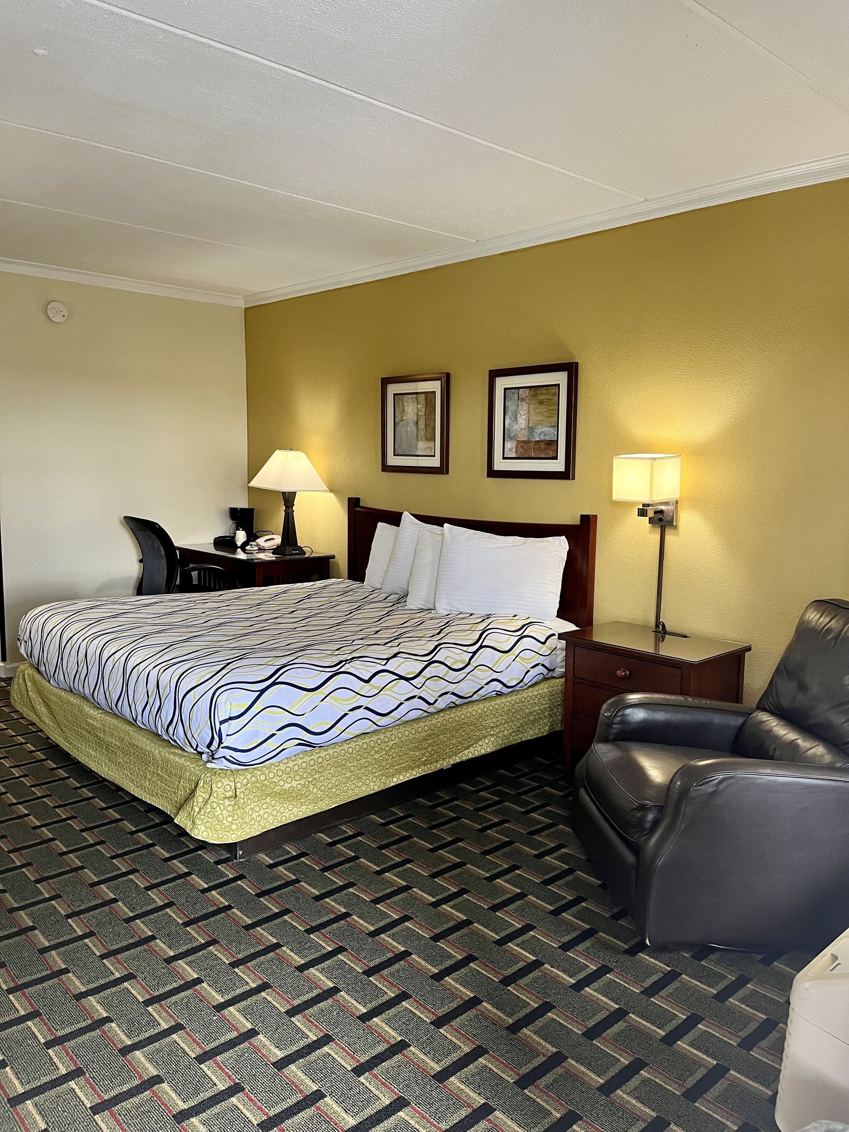 Ein Hotelzimmer mit einem Bett, einem Schreibtisch, einem Stuhl, zwei Wandleuchten und einem gemusterten Teppich.
