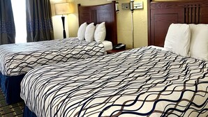 A hotel room with two beds, a headboard, a bedside table with a lamp, a framed picture on the wall, and a window with curtains. - West Bank Inn (Augusta)