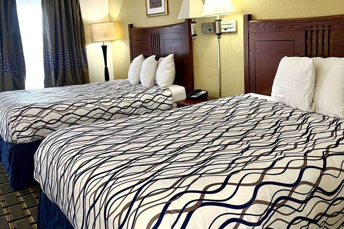 A hotel room with two beds, a headboard, a bedside table with a lamp, a framed picture on the wall, and a window with curtains.