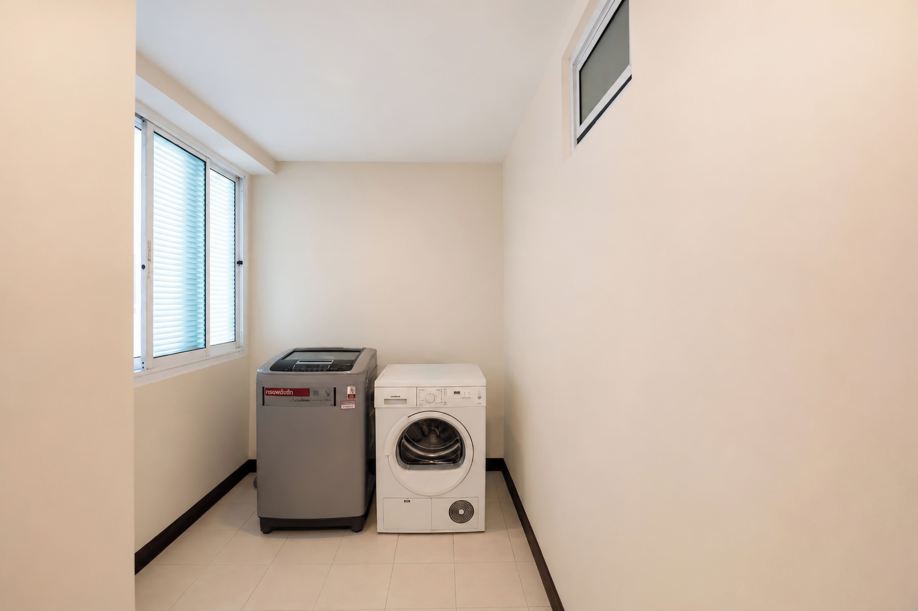 A laundry room with a washing machine and a dryer, a window with blinds, and a tiled floor.