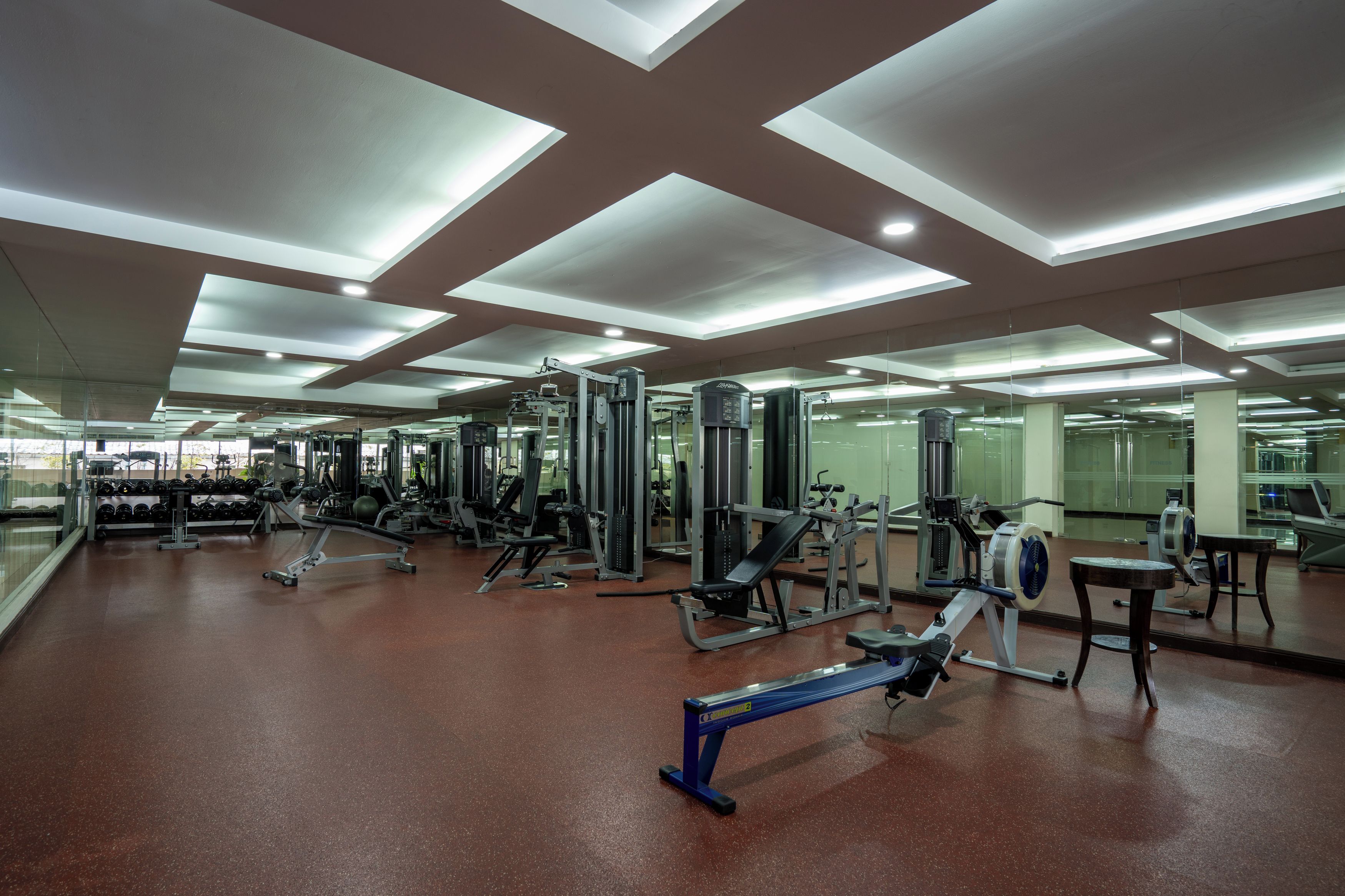 Une salle de sport équipée de divers appareils d’exercice, dotée de murs couverts de miroirs et d’un sol marron.