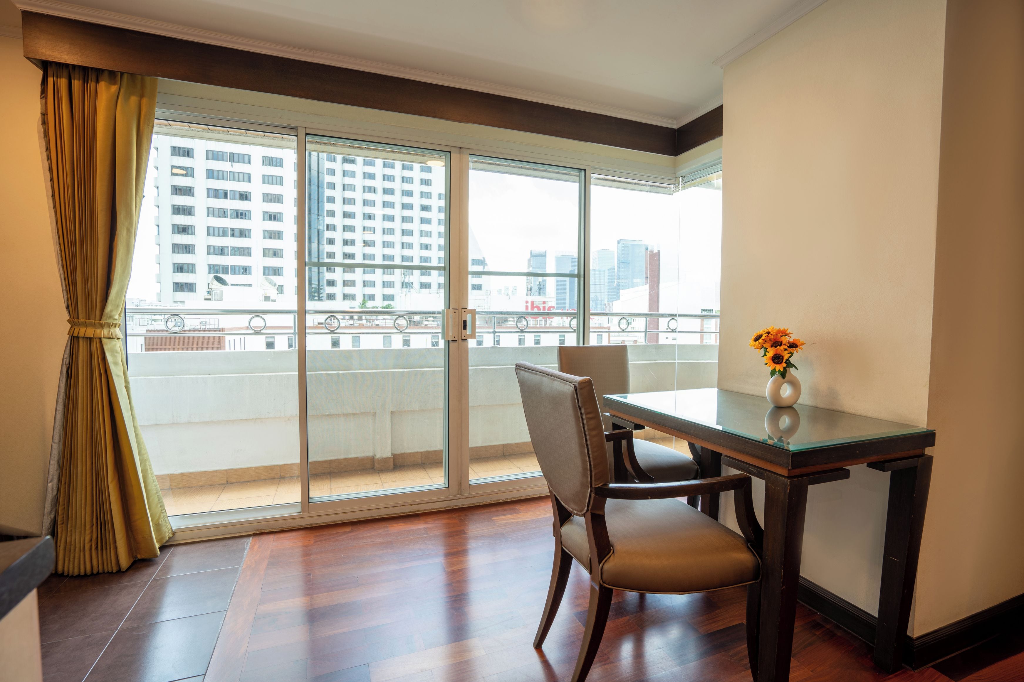 A room with a dining table, chairs, a vase with flowers, a window with curtains, and a view of buildings.