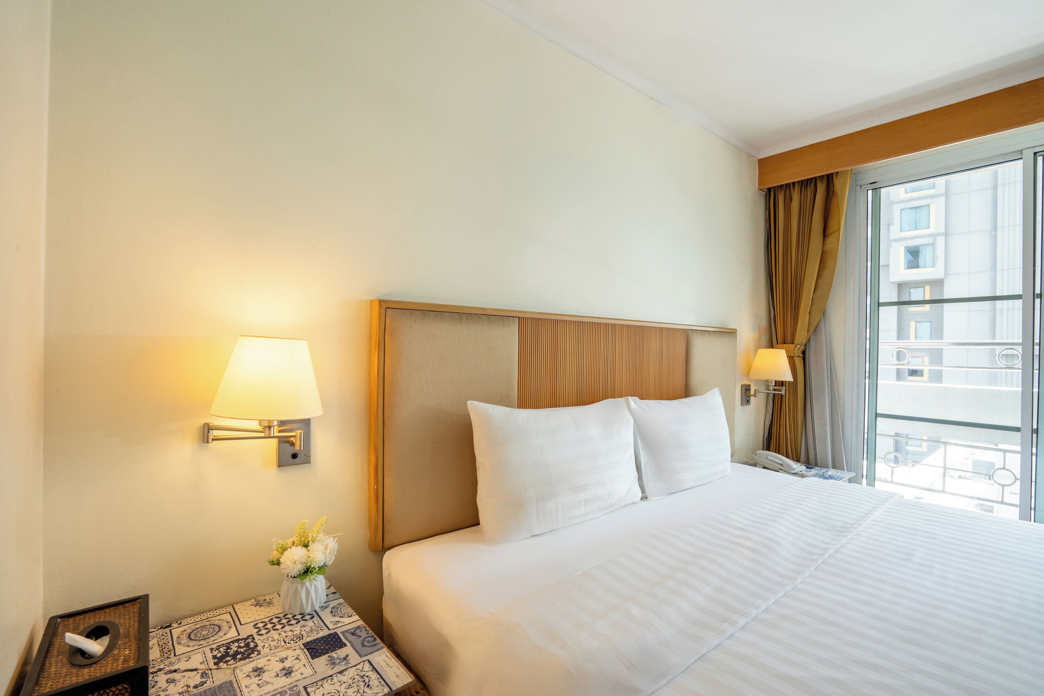 Quarto de hotel com uma cama grande, duas luminárias de cabeceira, mesa de cabeceira com um vaso de flores e vista para edifícios pela janela.