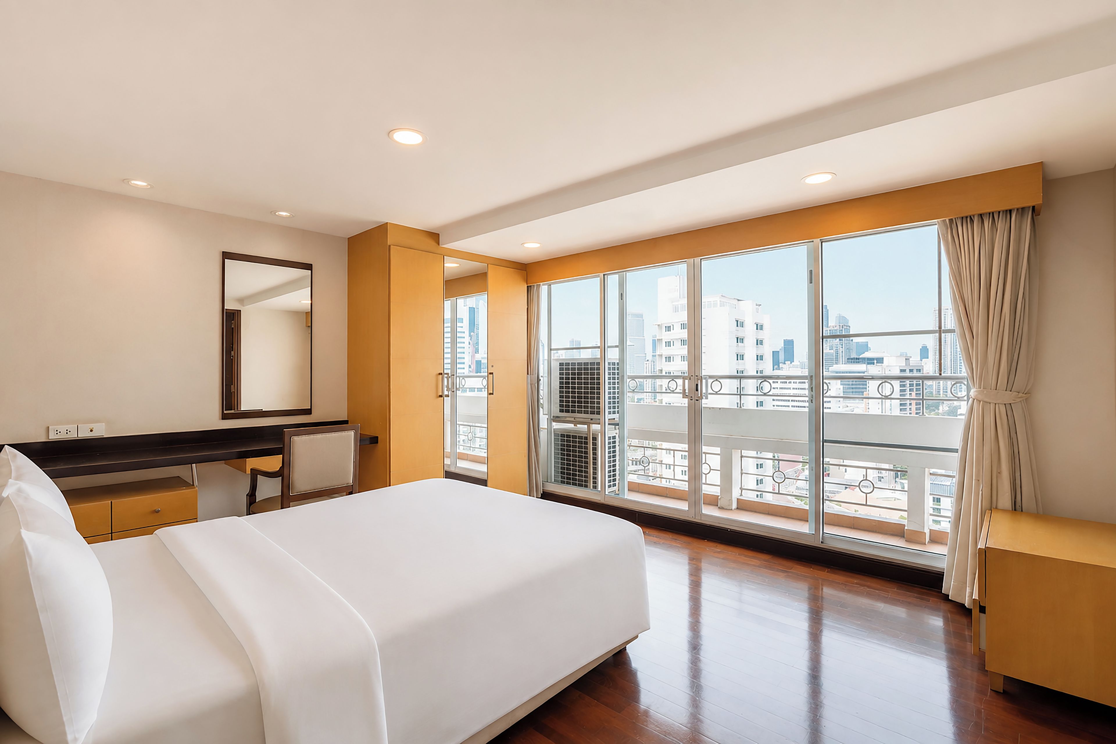 A hotel room with a large bed, a desk, and a view of the city through floor-to-ceiling windows.