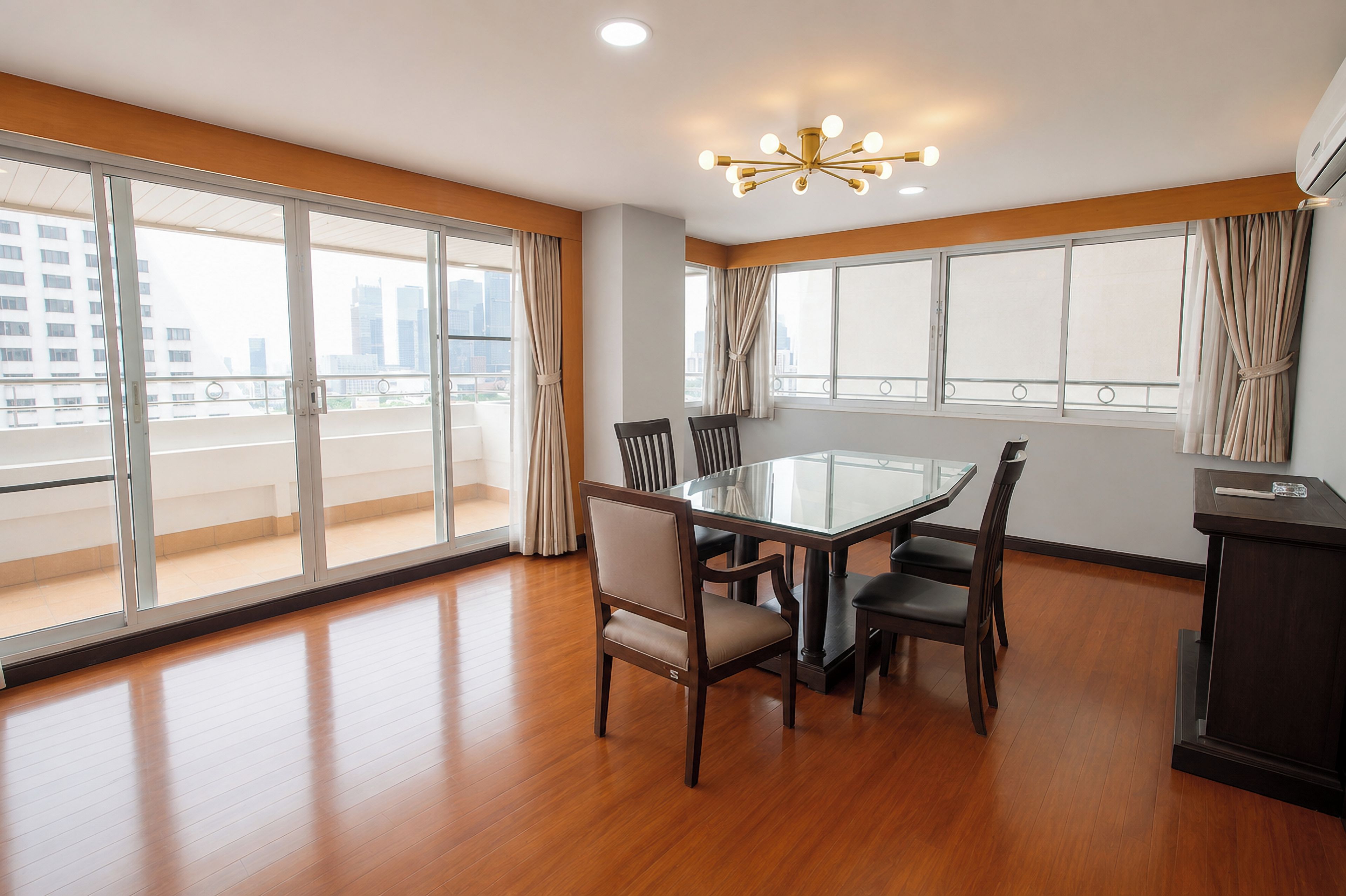 A modern dining area with a glass table, wooden chairs, and a city view through large windows.