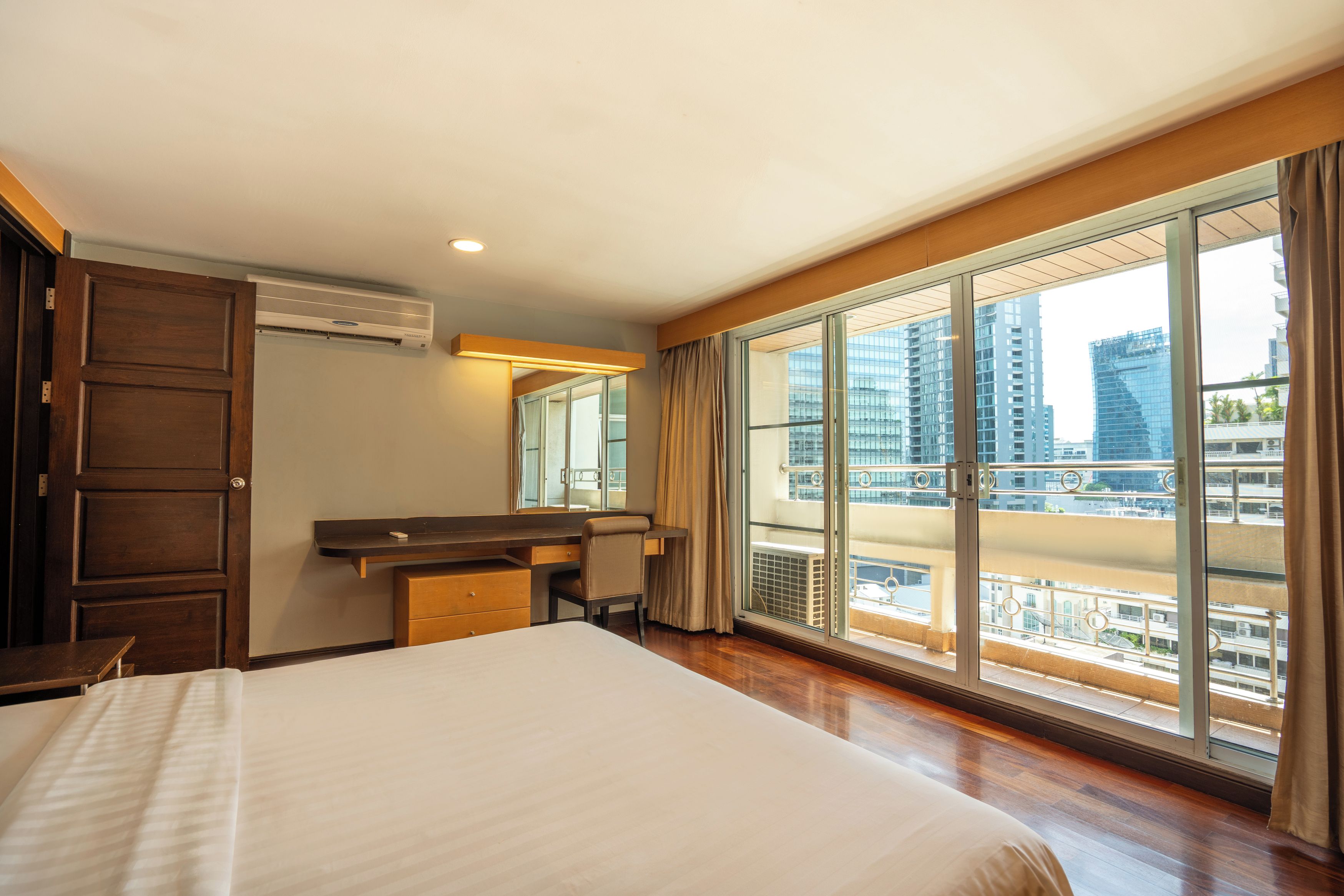 A hotel room with a large bed, a desk, a chair, and a view of the cityscape through a large window.