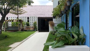 A modern building with a tiled roof, a pathway leading to a gated entrance, and lush greenery including large plants and trees. - Tree Residences (Chiang Mai)