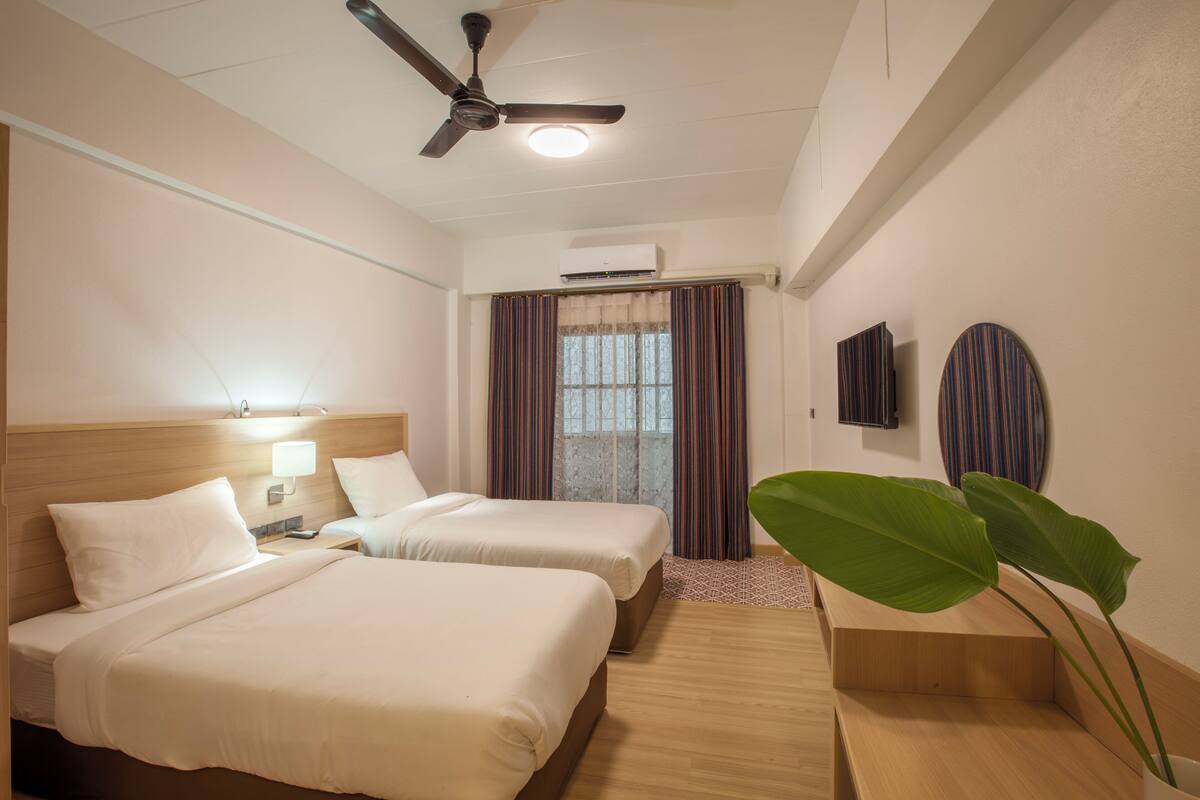 A hotel room with two beds, a ceiling fan, an air conditioning unit, a window with curtains, a bedside table with a lamp, and a large plant.